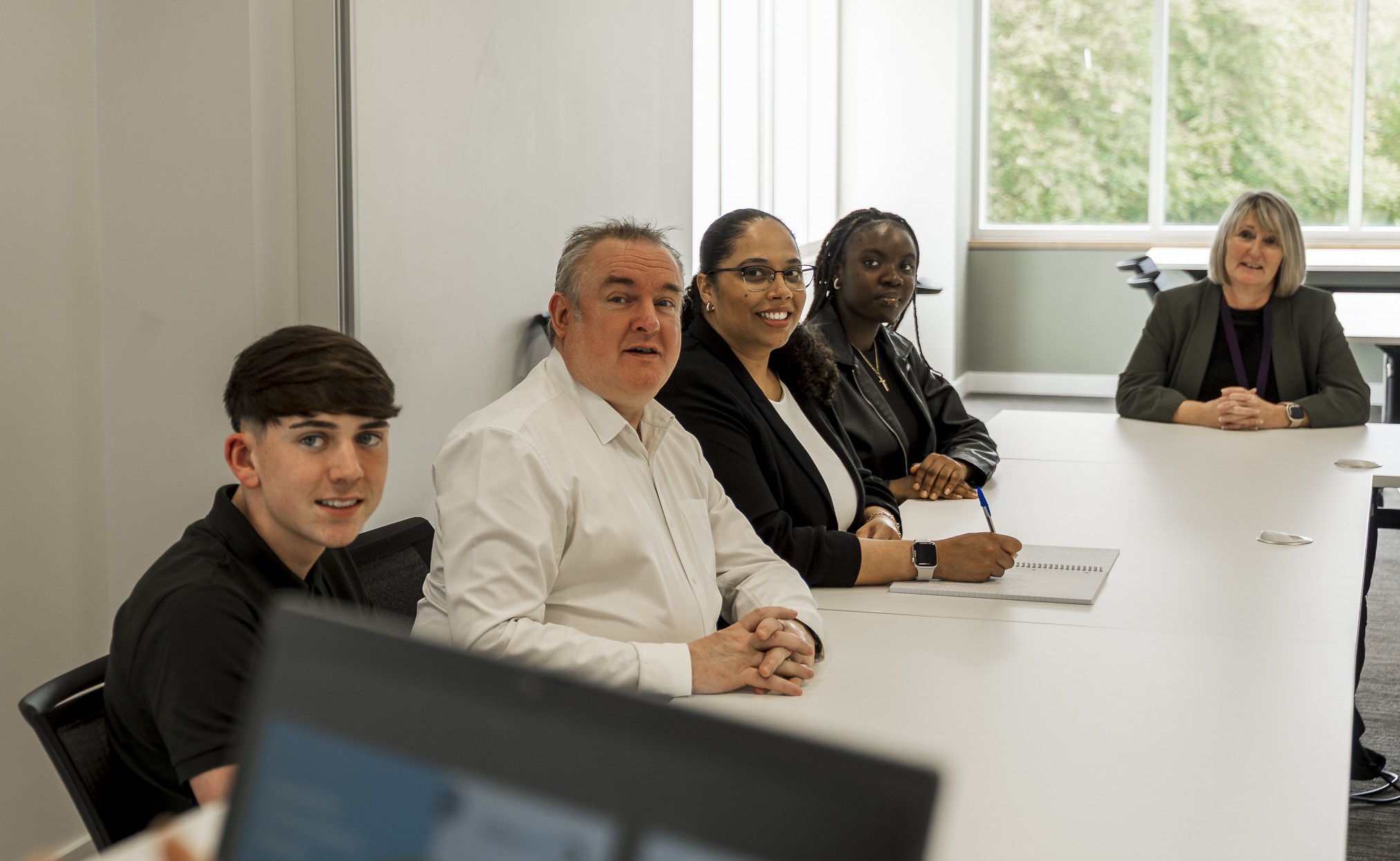 Three women and two men seated around a large conference table listening to someone off screen