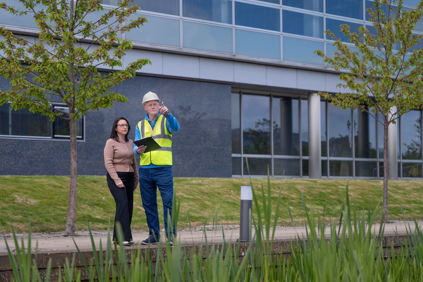 Long shot of man in hard hat with woman. Pond in foreground, office building in background.