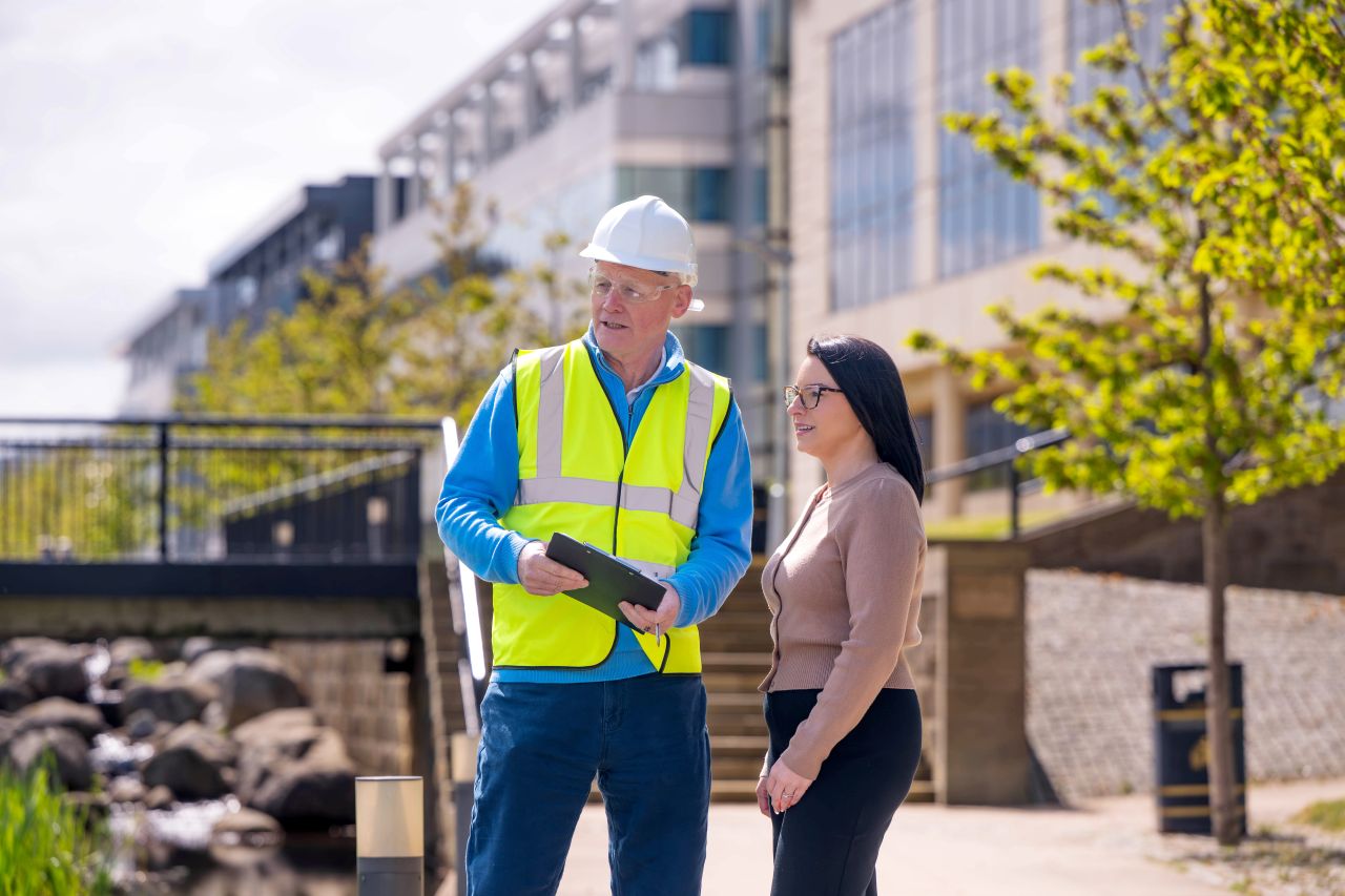 Man in hard hat and hi-vis jacket speaking to woman outside with buildings and landscaping in background
