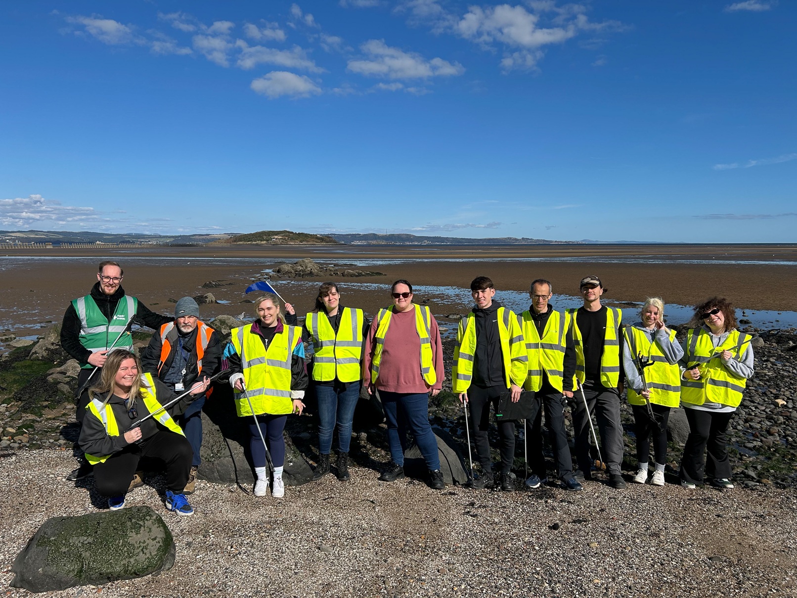A group of 11 smiling people on a beach, wearing hi-vis vests and holding litter pickers