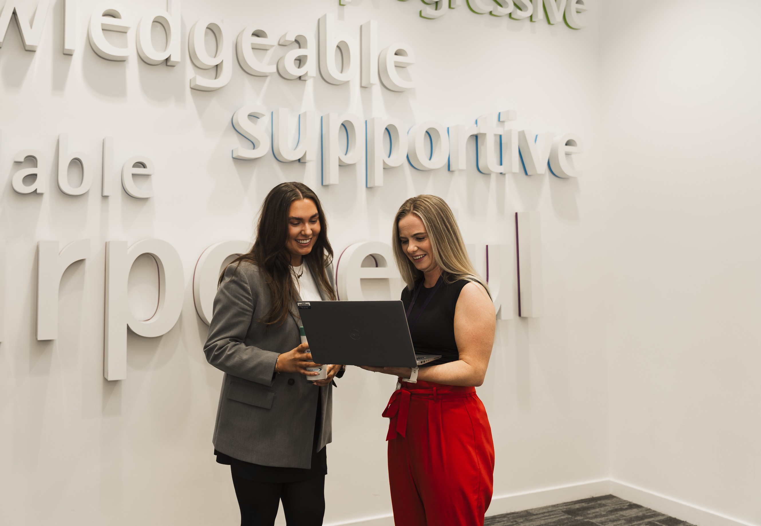 Two smiling women standing in front of a sign with the word "supportive" visible while looking at a laptop