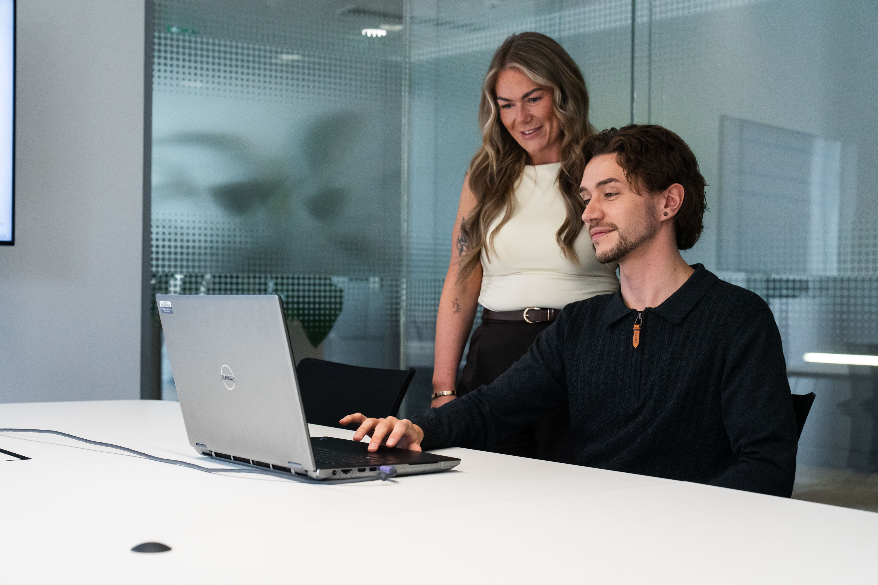 Woman standing behind man seated as both look at laptop screen with glass partition office behind them