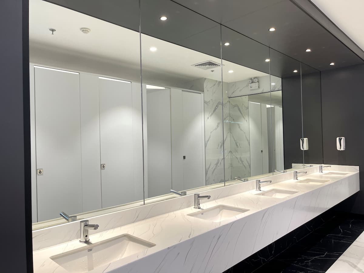 Row of modern white sinks in public toilet with cubicles reflected in mirror wall above
