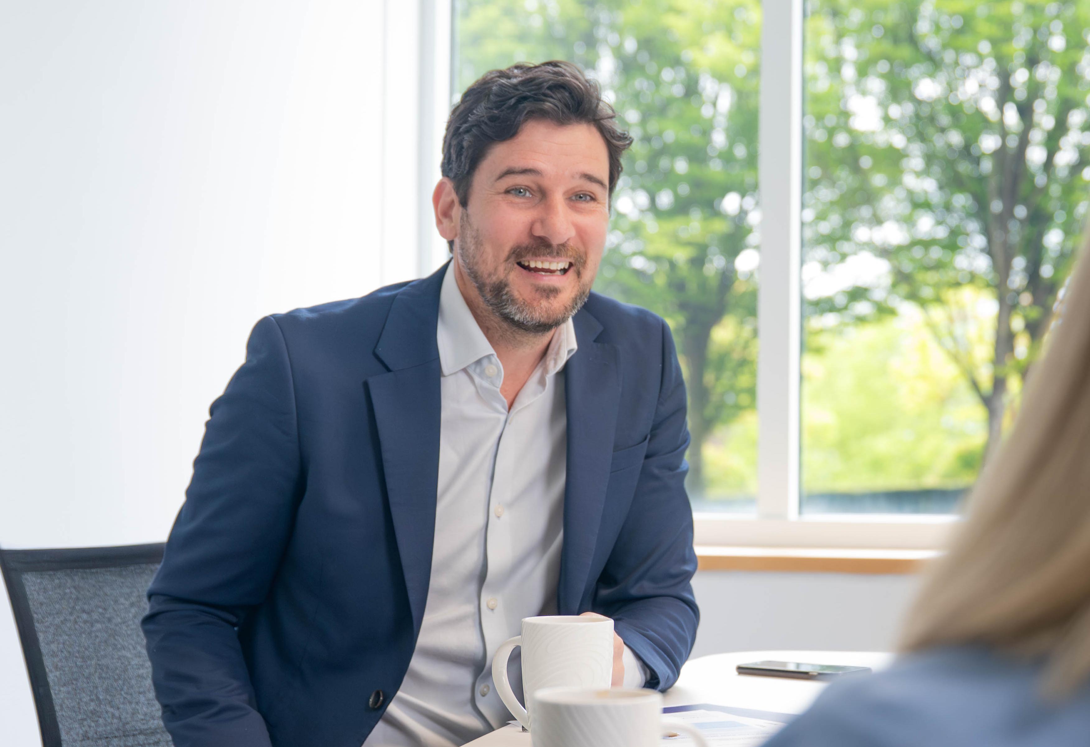 Chris Arbuckle in suit with no seated at a meeting table, smiling and talking