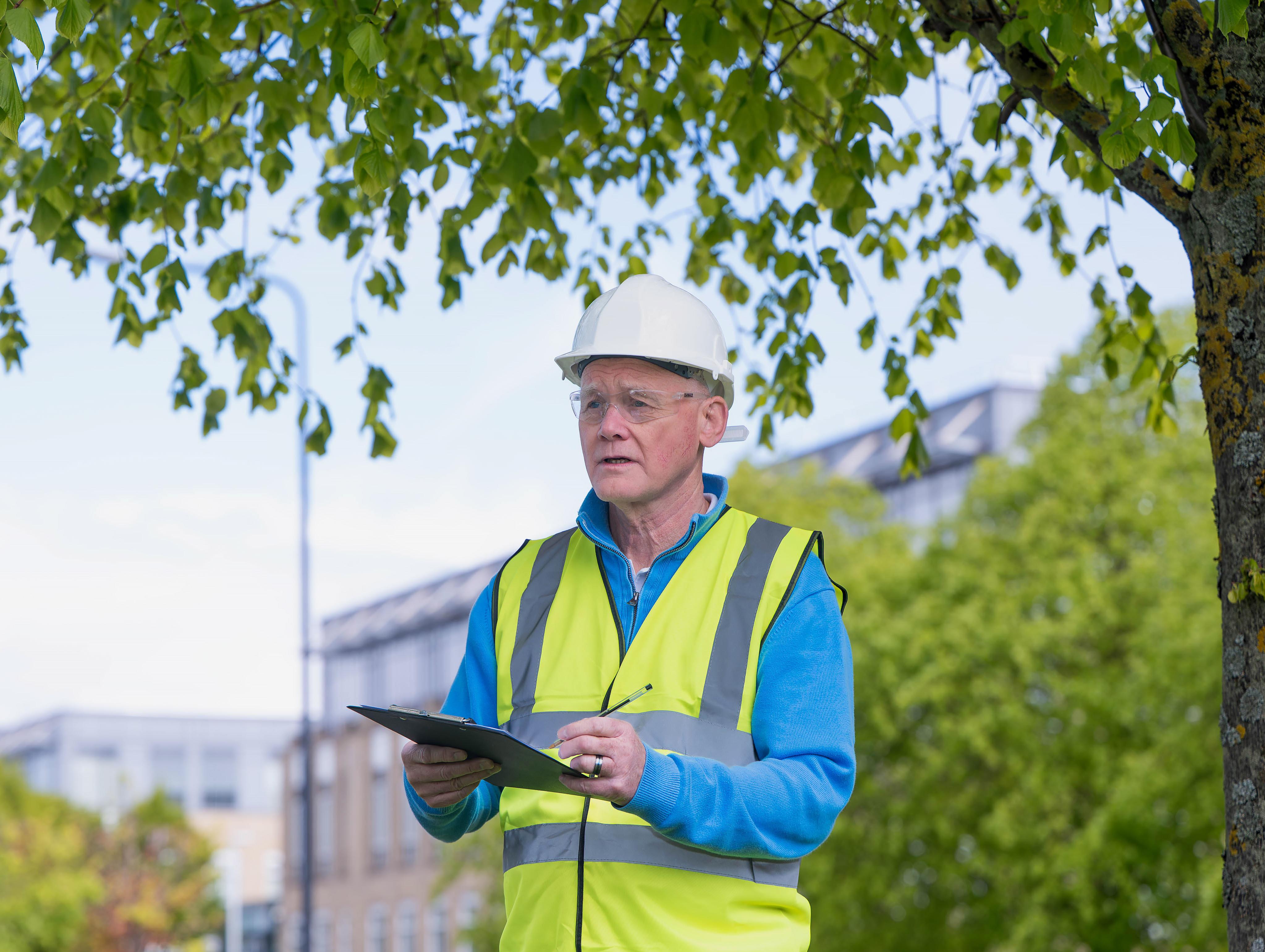Man in hard hat holding clipboard with office building and trees in background