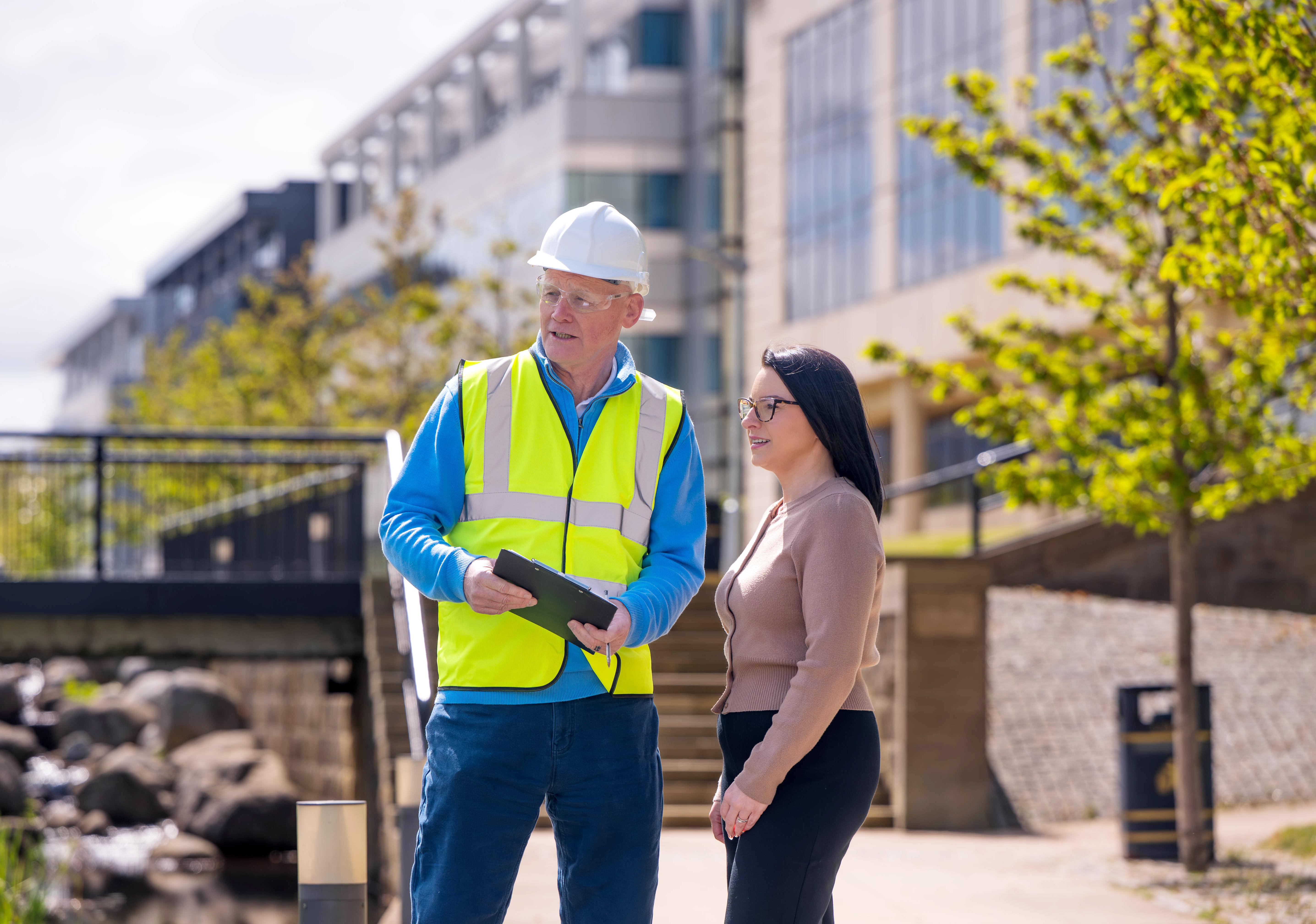 Man in hard hat and hi-vis jacket speaking to woman outside with buildings and landscaping in background