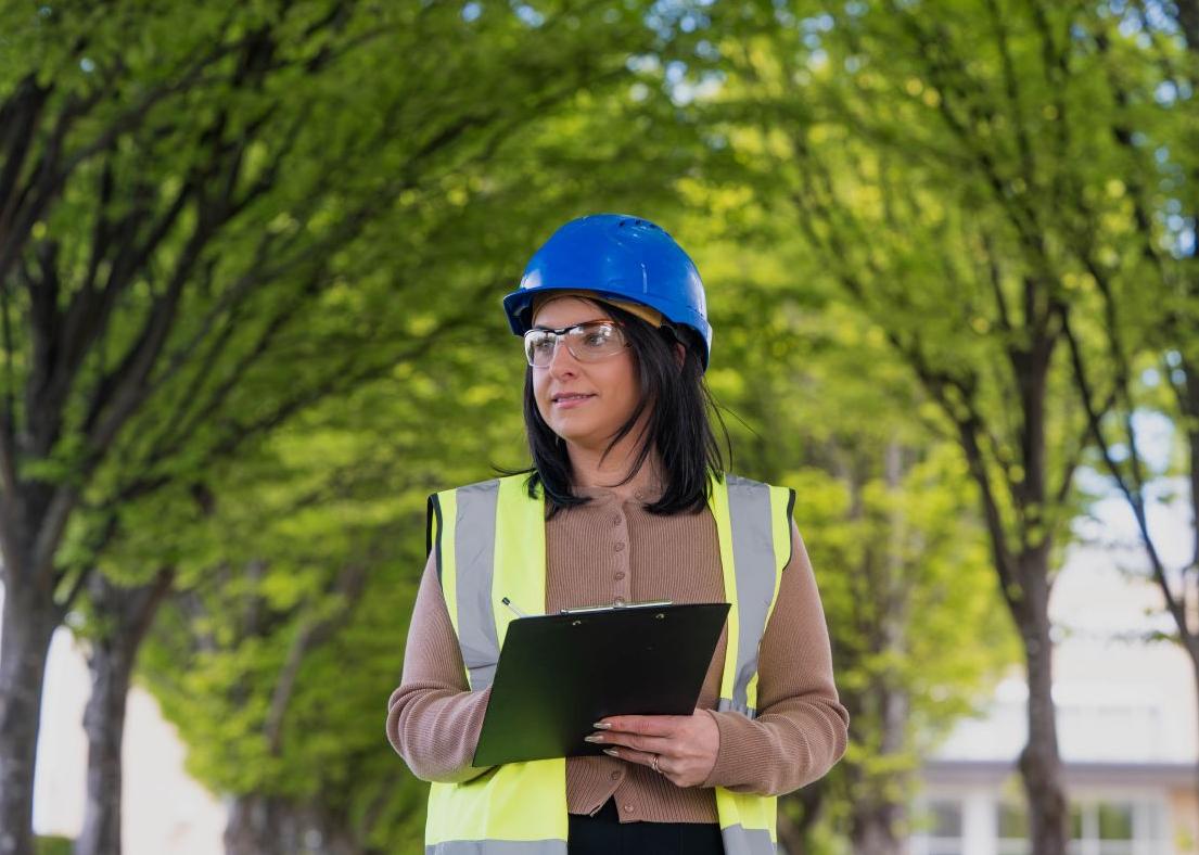 Woman in blue hard hat holding clipboard with green trees in background