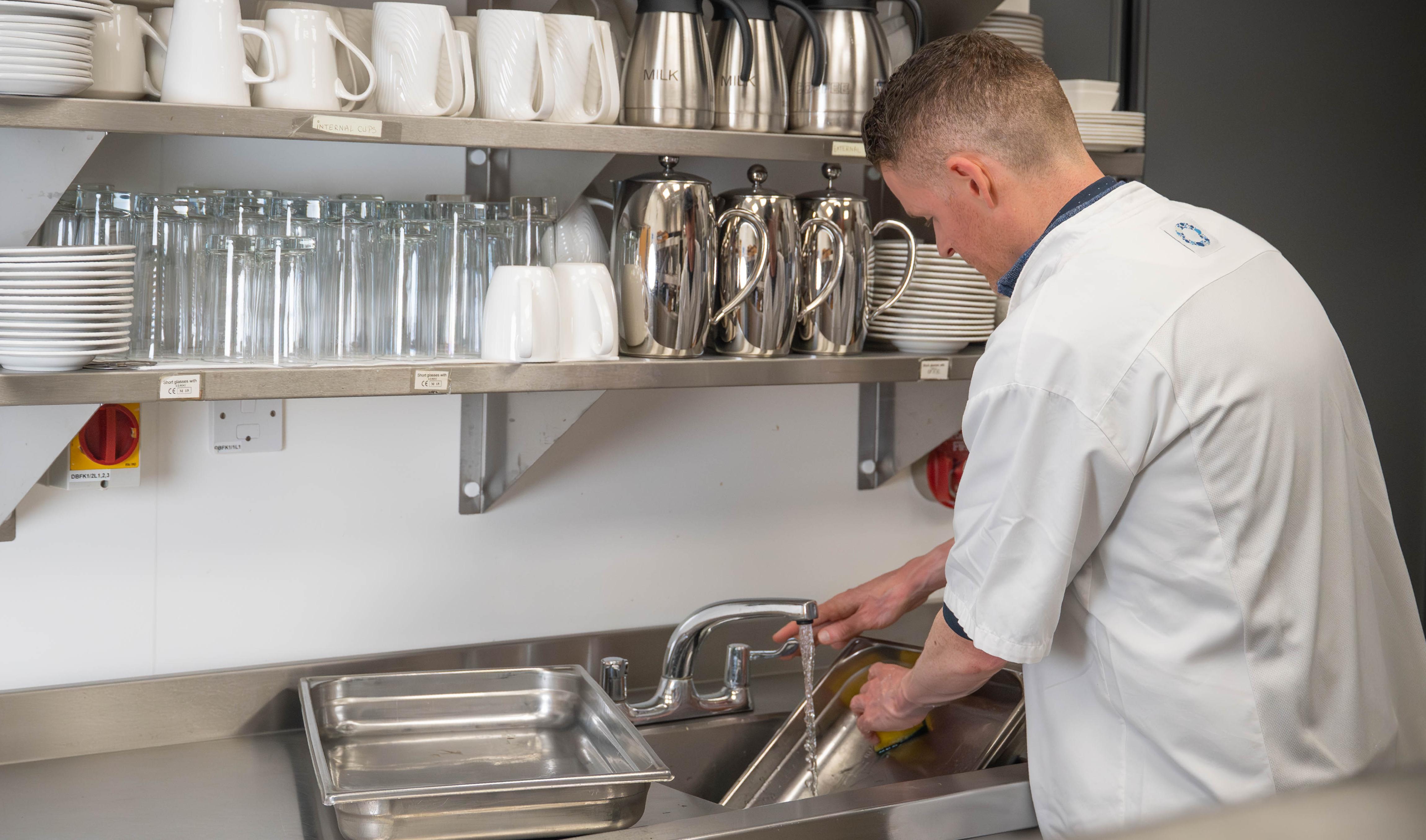 Man in catering overalls viewed from behind washing equipment at a commercial sink with shelving above full of cups, plates, glasses etc.