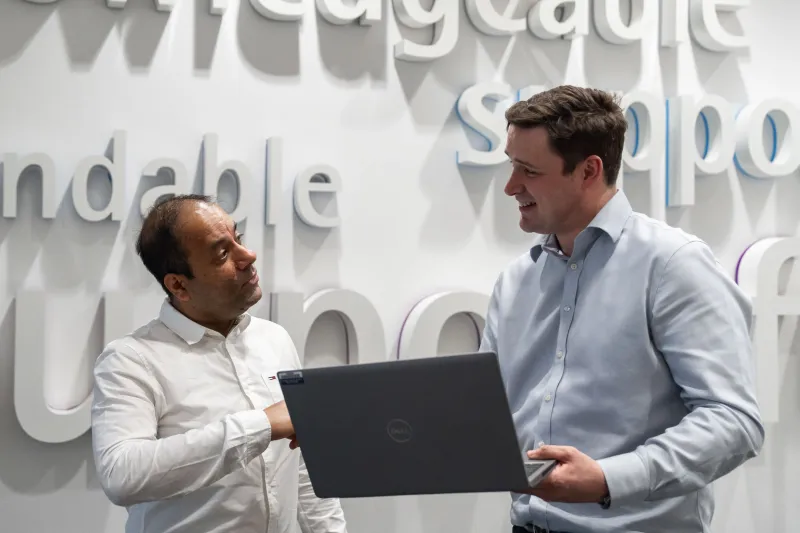 Two men standing in front of a word wall talking and looking at a laptop