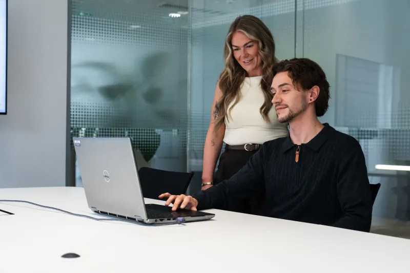 Woman standing behind man seated as both look at laptop screen with glass partition office behind them